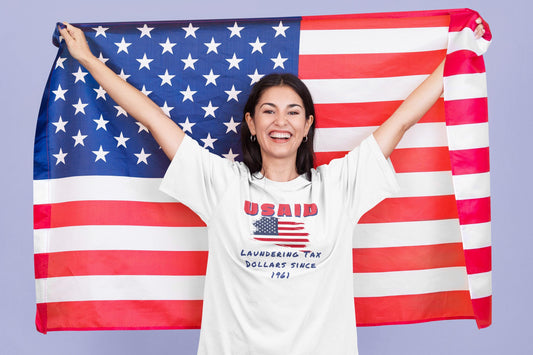 A smiling woman holds up a large American flag behind her, standing against a light purple background. She is wearing a white T-shirt featuring a distressed American flag graphic and bold red text that reads "USAID." Below the flag, the shirt says in blue handwritten-style text: "Laundering Tax Dollars Since 1961." The overall tone of the shirt is satirical and politically charged.