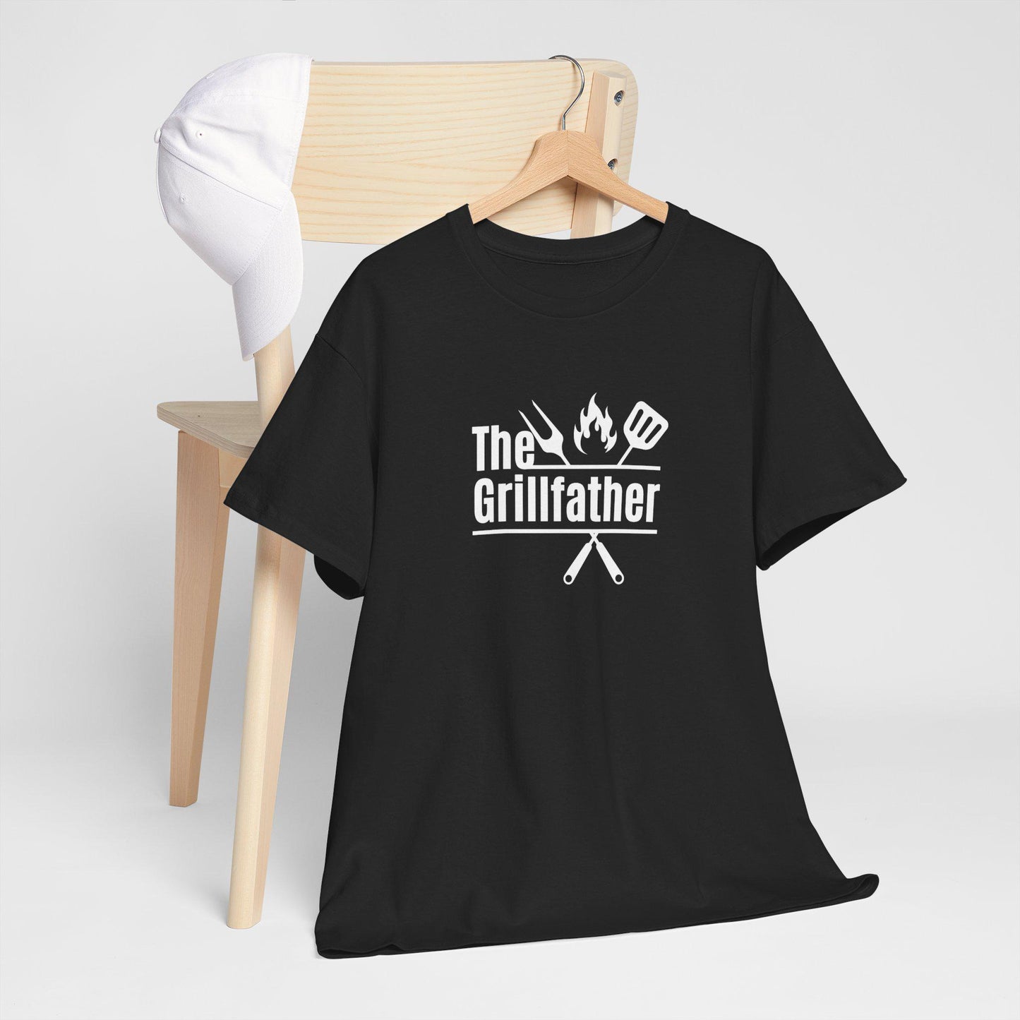 Black T-shirt draped over a wooden chair with a white cap on the backrest, featuring the phrase “The Grillfather” in white text with grilling icons of a fork, flame, and spatula above crossed utensils.