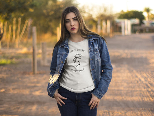 A young woman stands on a dirt road during golden hour, wearing a light-colored graphic t-shirt with a line drawing of a rooster's head and the text "CLUCK AROUND AND FIND OUT." She has long straight hair and a serious expression. Over the t-shirt, she wears a blue denim jacket paired with dark jeans. The background features trees, fencing, and a warm, softly lit rural setting.