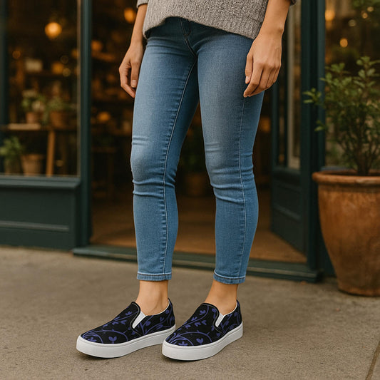 Person wearing slip-on sneakers with a black base and dark purple ivy vine pattern, standing outside a cozy storefront. The shoes are paired with fitted blue jeans and a gray sweater, creating a casual and stylish fall outfit.
