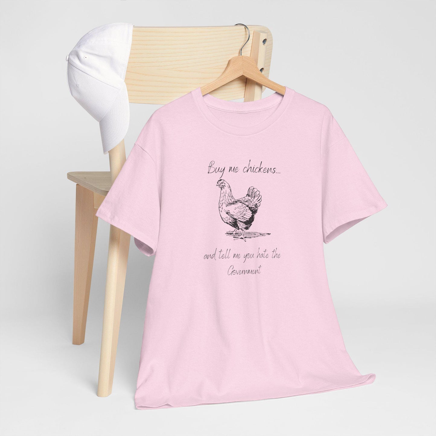 A light pink T-shirt hangs on a wooden hanger over the back of a light wood chair, with a white baseball cap resting on the chair. The T-shirt features a black sketch-style illustration of a chicken and the playful text: "Buy me chickens... and tell me you hate the Government." The minimalist setup emphasizes the shirt's quirky message and soft pastel color.