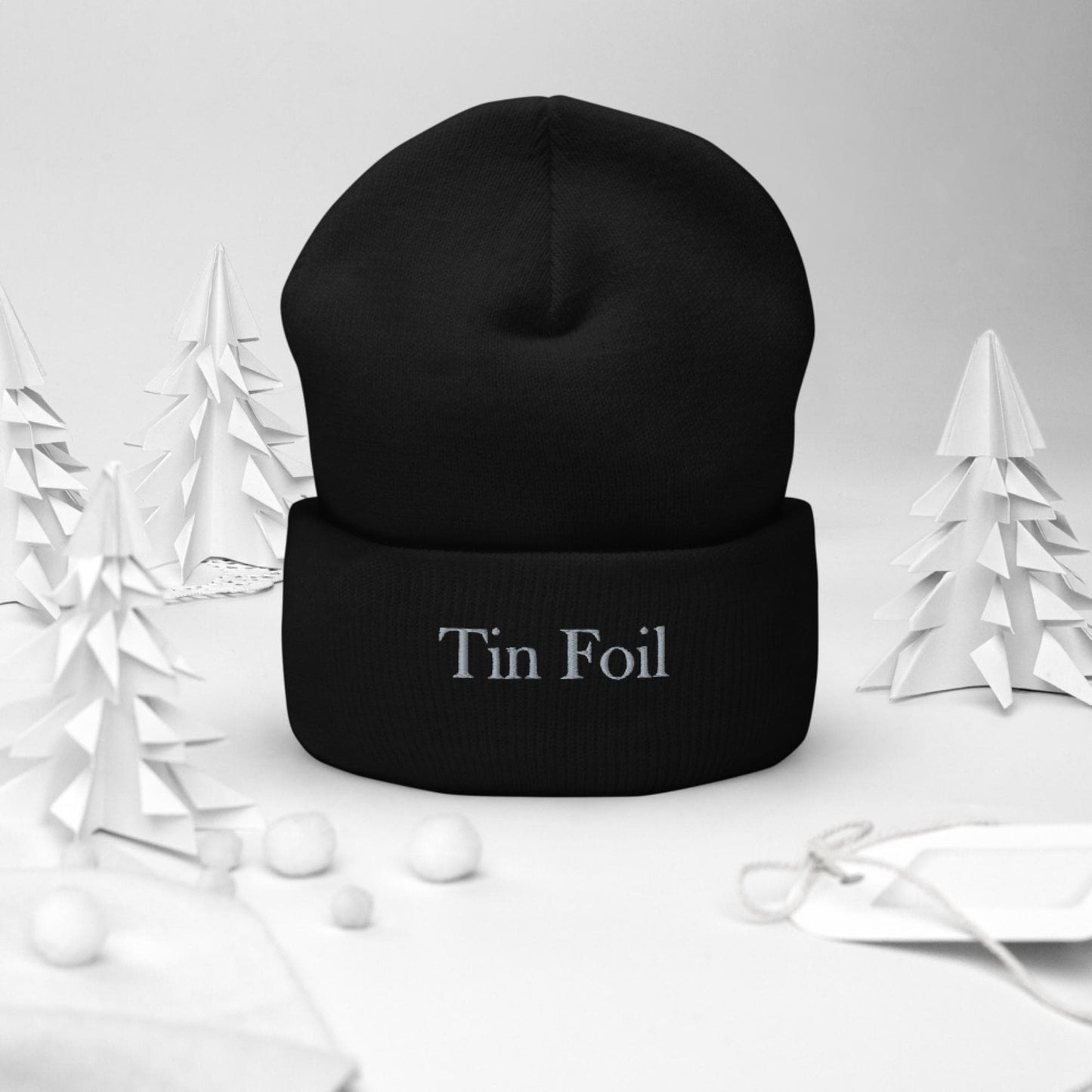 "Black beanie with the words 'Tin Foil' embroidered on the front, displayed upright in a winter-themed setting. Surrounding the beanie are white paper-cut trees, small white pom-pom snowballs, and a white gift tag with string, all set against a soft white background."