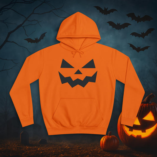 Bright orange hoodie with a black jack-o'-lantern face design on the front, laid flat against a spooky Halloween-themed background. The setting includes a foggy night sky, flying bats, bare tree branches, and glowing jack-o'-lanterns on the ground near gravestones.