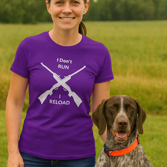Woman standing in a grassy outdoor field with a brown and white hunting dog wearing an orange collar. She is smiling and wearing a purple T-shirt featuring two crossed rifles and the text “I Don’t RUN I RELOAD” printed in white.