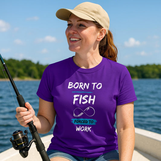 Smiling woman sitting in a boat on the water, holding a fishing rod. She is wearing a beige cap and a purple T-shirt with the text “BORN TO FISH, FORCED TO WORK” and a graphic of a fishing hook shaped like an infinity symbol. Trees and blue sky are visible in the background.