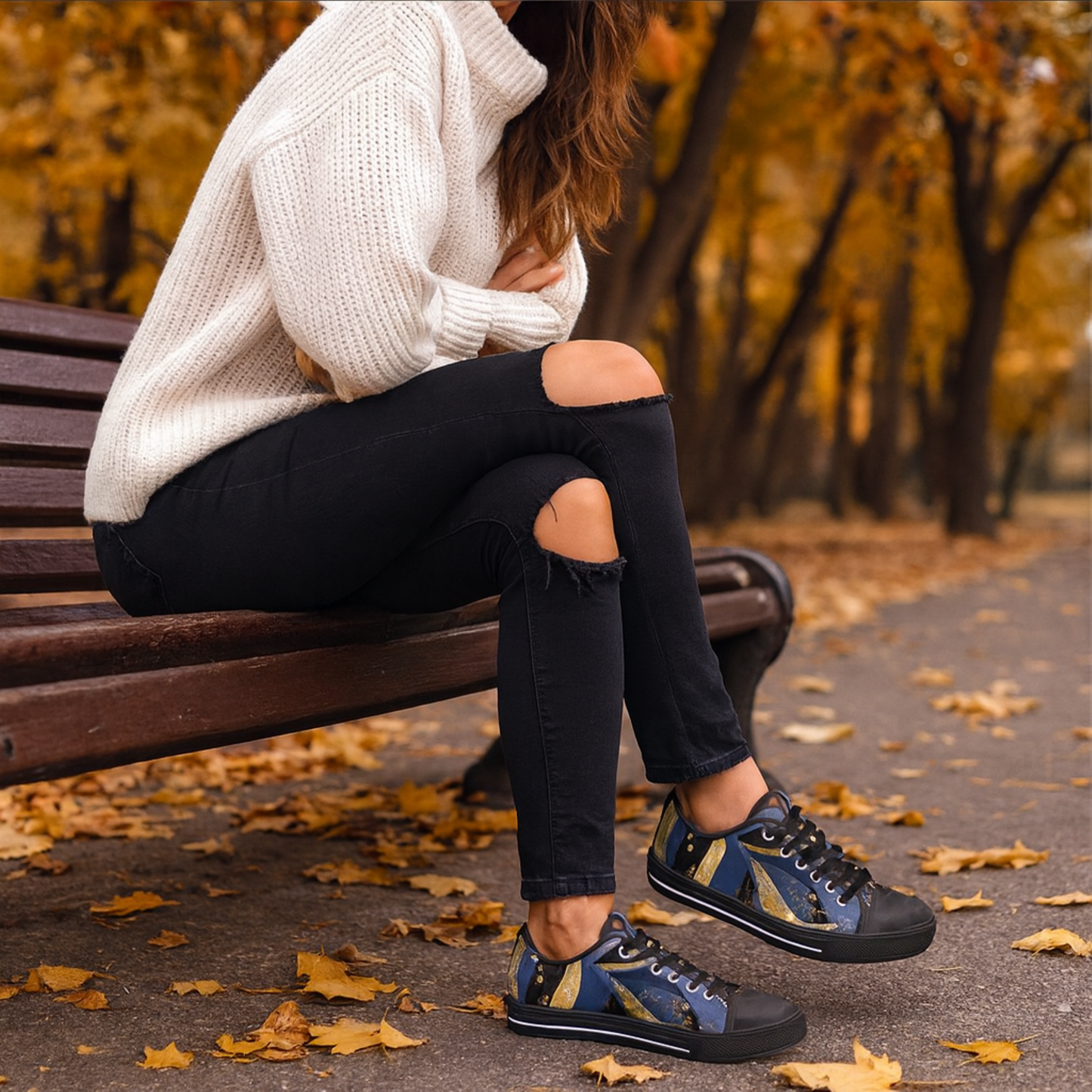 A woman sitting on a bench wearing a white sweater and black ripped jeans with blue & gold abstract patterned sneakers in an autumn setting.