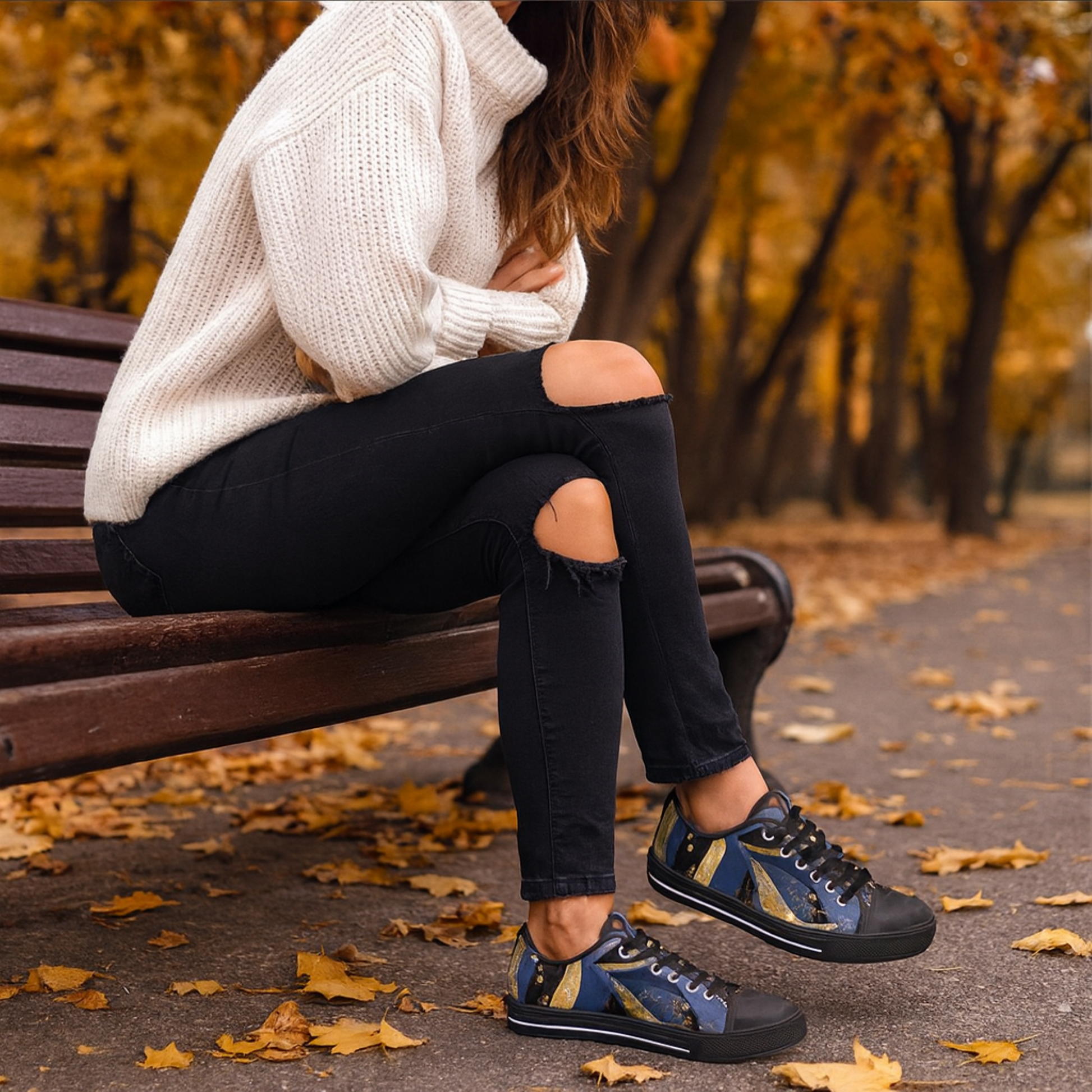 A woman sitting on a bench wearing a white sweater and black ripped jeans with blue & gold abstract patterned sneakers in an autumn setting.