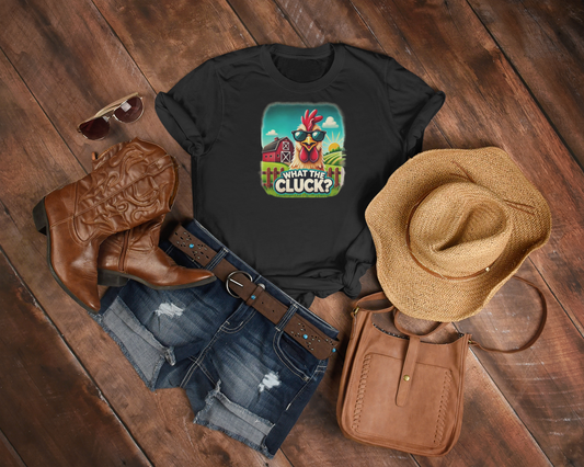 Flat lay of a country-inspired outfit featuring a black t-shirt with a colorful cartoon chicken wearing sunglasses and the phrase "What the Cluck?" set against a farm background. The ensemble includes distressed denim shorts, a brown leather belt, cowboy boots, a straw hat, a brown crossbody bag, and aviator sunglasses, all arranged on a rustic wooden floor.