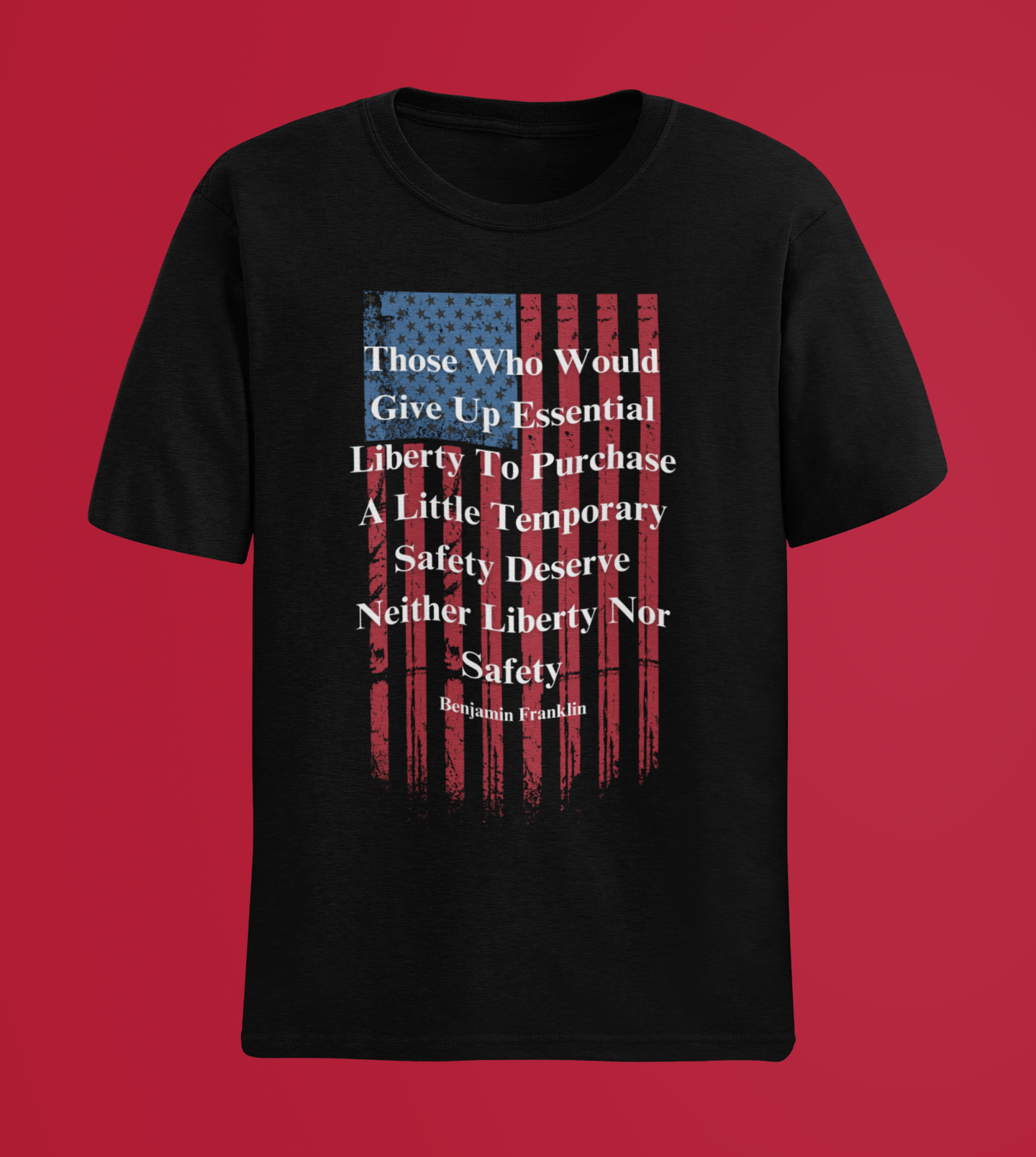 A black T-shirt is displayed against a solid red background. The shirt features a distressed vertical American flag graphic in red and blue, overlaid with a bold white quote by Benjamin Franklin: “Those who would give up essential liberty to purchase a little temporary safety deserve neither liberty nor safety.” The striking patriotic design stands out prominently against the red backdrop, emphasizing freedom and American values.