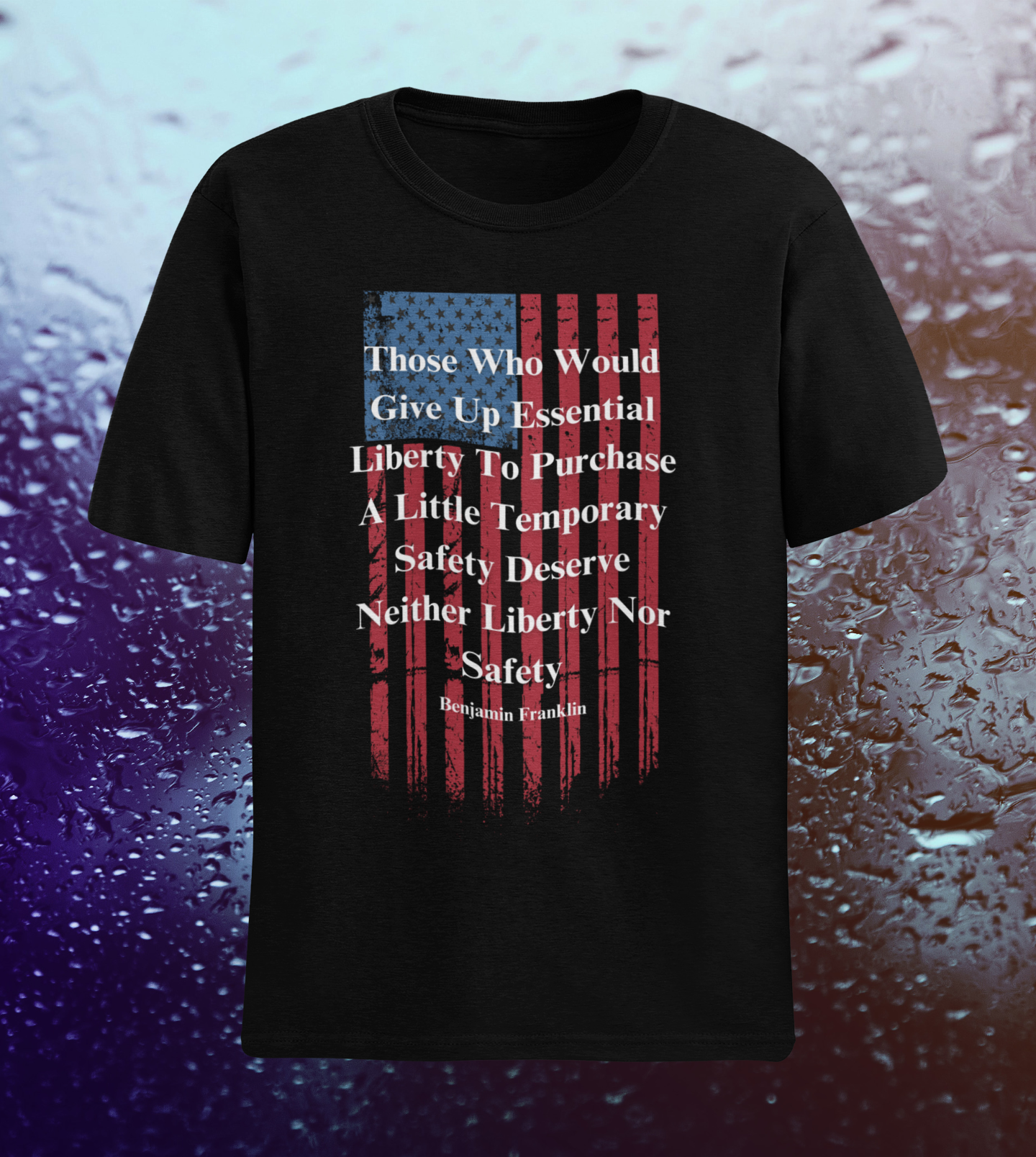 A black T-shirt is displayed against a backdrop of rain-covered glass with a gradient of blue and purple tones. The shirt features a distressed vertical American flag design in red and blue, with a bold white quote by Benjamin Franklin printed over it: “Those who would give up essential liberty to purchase a little temporary safety deserve neither liberty nor safety.” The shirt's patriotic and bold design stands out against the moody, weathered background.