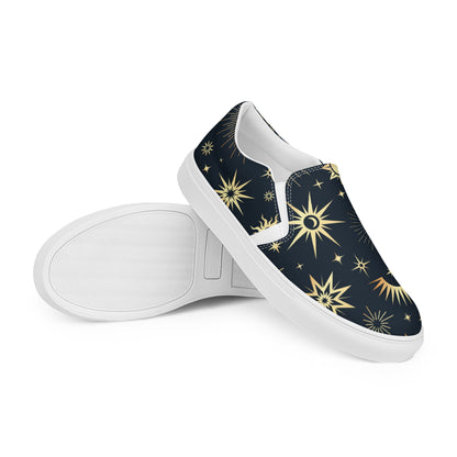 Pair of slip-on canvas shoes featuring a dark navy background with a gold celestial pattern of suns, stars, and crescent moons. One shoe is positioned upright while the other is laid on its side to show the textured white rubber sole. Displayed on a white background.