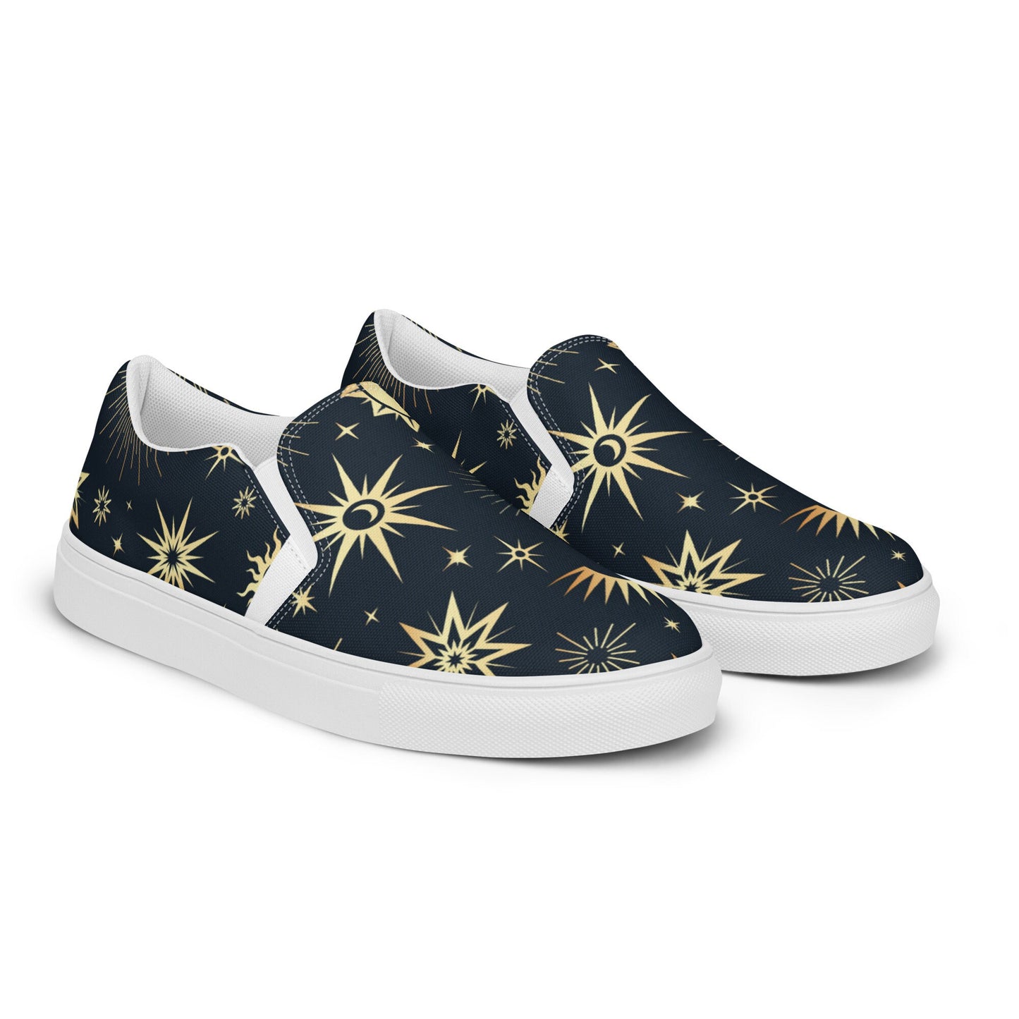 Pair of slip-on canvas shoes with a navy blue background and an all-over celestial print featuring gold and yellow stars, suns, and crescent moons. The shoes have white soles and inner lining, displayed on a clean white background.
