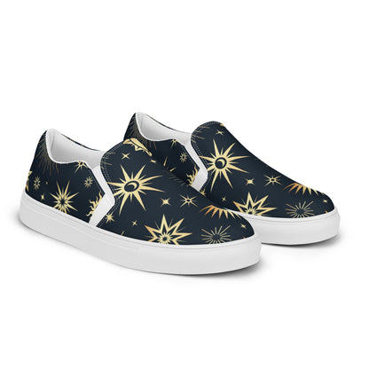 Pair of slip-on canvas shoes with a navy blue background and an all-over celestial print featuring gold and yellow stars, suns, and crescent moons. The shoes have white soles and inner lining, displayed on a clean white background.