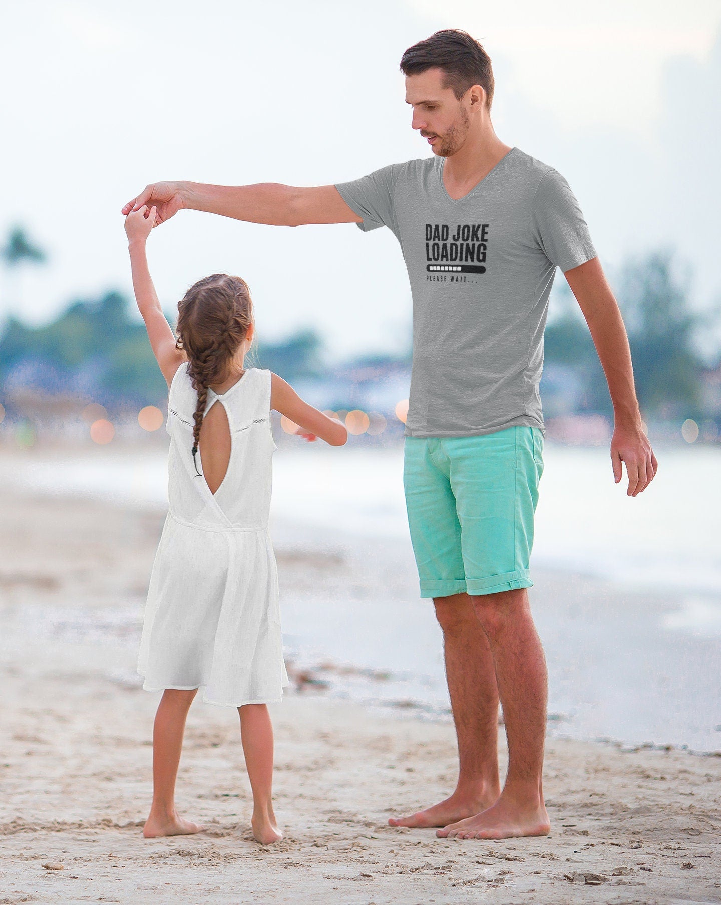 Father in a light gray “Dad Joke Loading, Please Wait…” T-shirt and mint green shorts dancing barefoot on the beach with his young daughter in a white dress, with the ocean and a soft-focus shoreline in the background.
