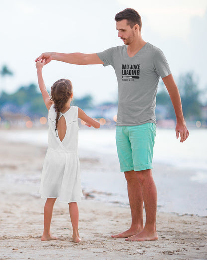 Father in a light gray “Dad Joke Loading, Please Wait…” T-shirt and mint green shorts dancing barefoot on the beach with his young daughter in a white dress, with the ocean and a soft-focus shoreline in the background.