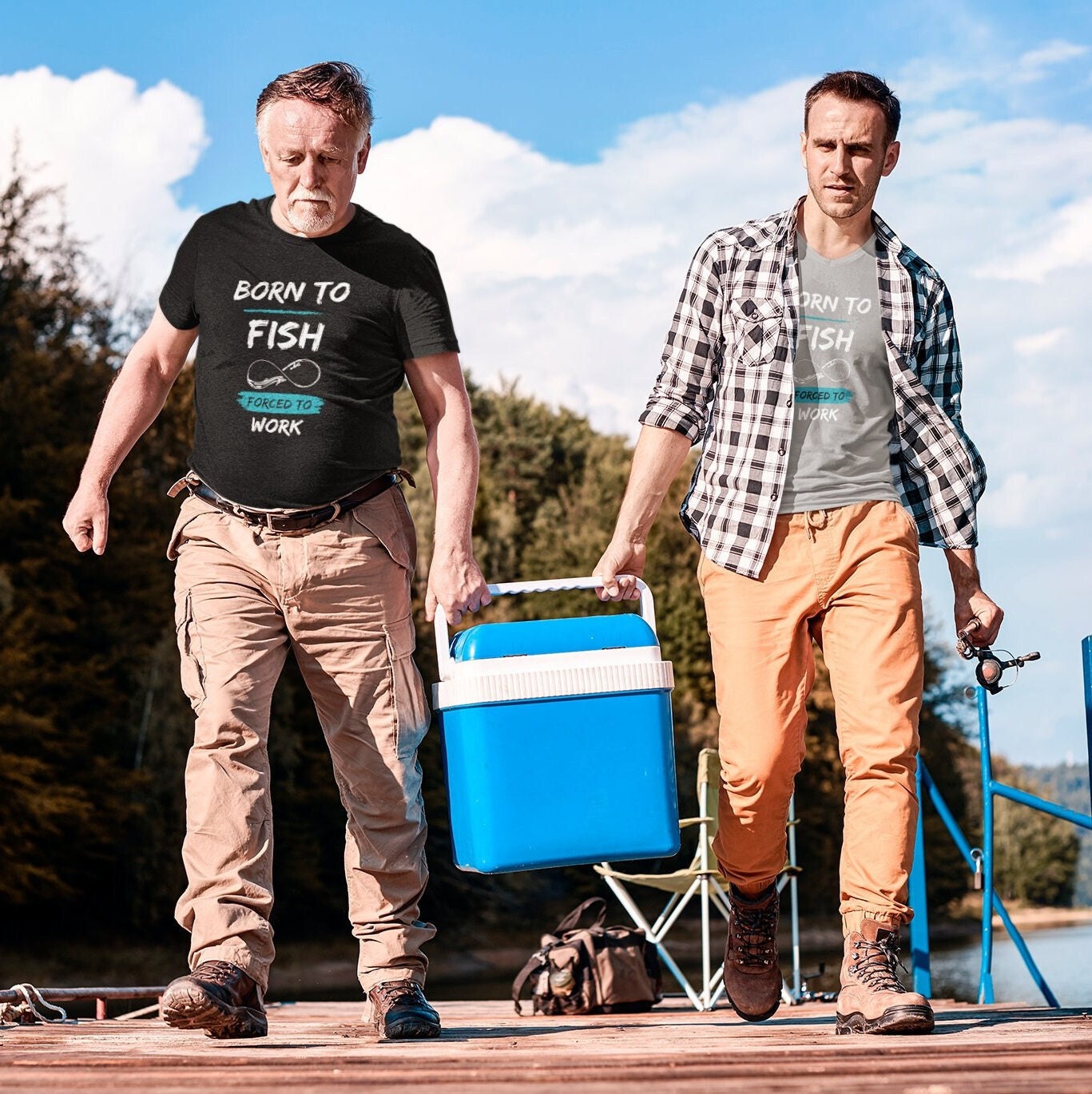 Two men walking on a wooden dock near a lake, carrying a large blue cooler together. Both are dressed in outdoor clothing and wearing T-shirts that say &quot;BORN TO FISH, FORCED TO WORK&quot; with a fish hook graphic. The older man on the left wears a black T-shirt and tan cargo pants, while the younger man on the right wears a light gray T-shirt, plaid shirt, and orange pants, holding a fishing rod. Trees and water are visible in the background under a partly cloudy sky.