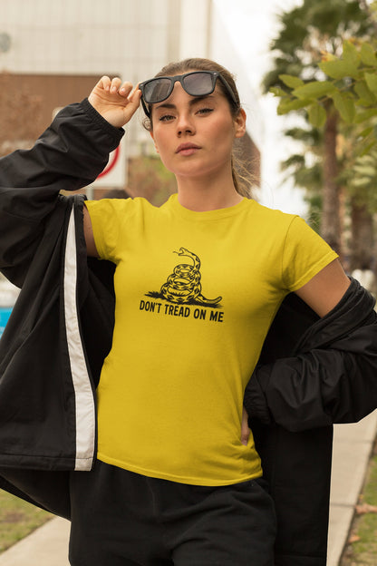 Woman wearing a yellow &quot;Don&#39;t Tread on Me&quot; t-shirt with a black coiled snake graphic, standing confidently outdoors with sunglasses and a black jacket draped over her shoulders. The background features palm trees and urban architecture.