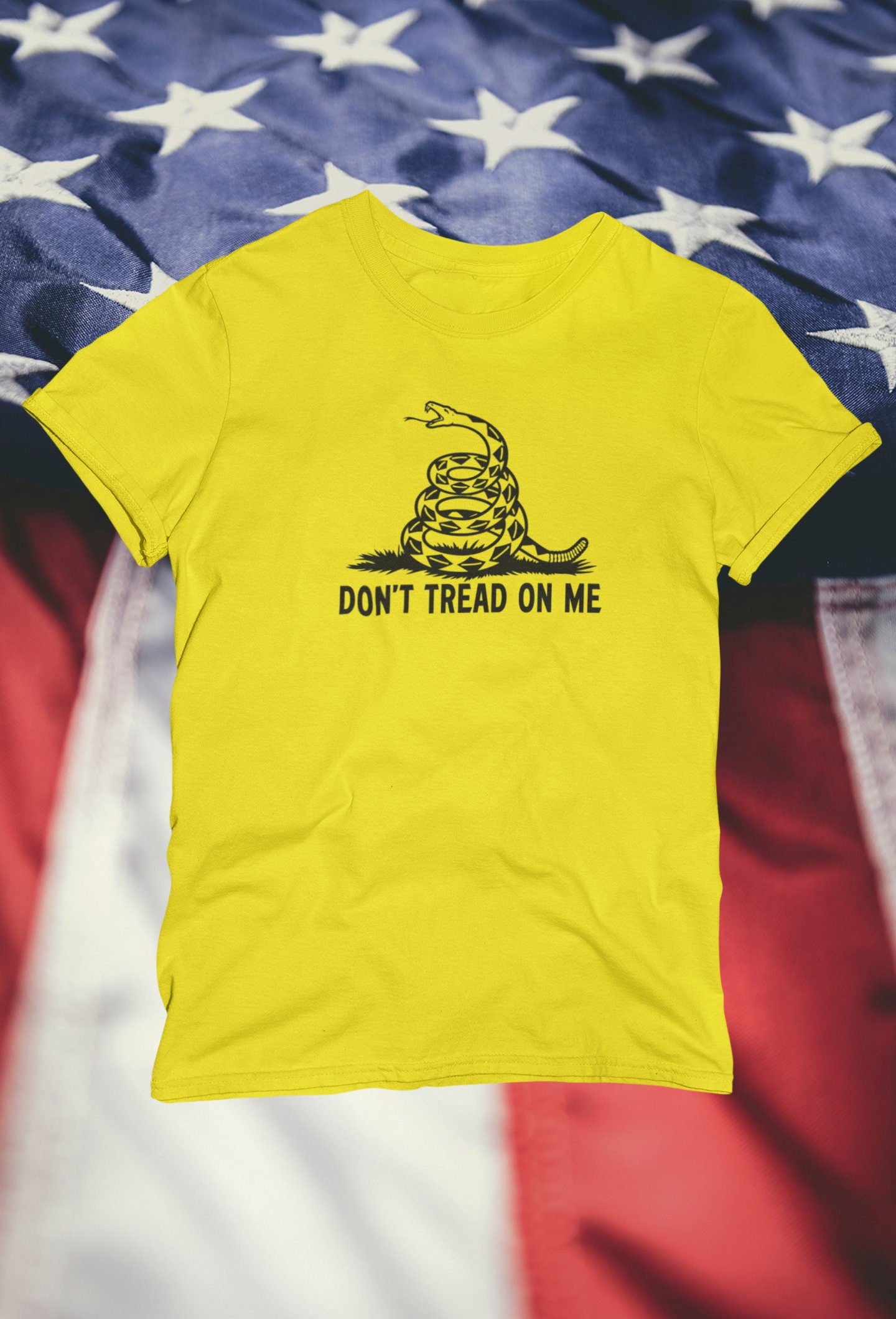 Bright yellow t-shirt with a black coiled snake graphic and the bold text &quot;DON&#39;T TREAD ON ME&quot; displayed against a blurred backdrop of the American flag featuring white stars on blue and red-and-white stripes.