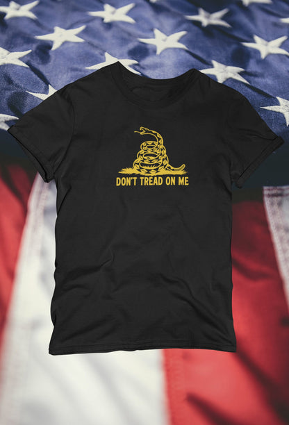 Black t-shirt featuring a yellow coiled snake graphic and the text &quot;DON&#39;T TREAD ON ME&quot; displayed against a blurred background of the American flag with white stars on blue and red-and-white stripes.