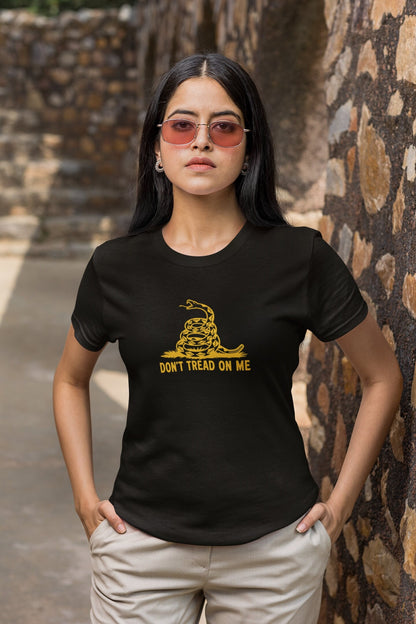 Woman wearing a black &quot;Don&#39;t Tread on Me&quot; t-shirt with a yellow coiled snake graphic, standing confidently outdoors with her hands in her pockets. She has long dark hair, is wearing pink-tinted sunglasses and earrings, and is positioned against a rustic stone wall background.