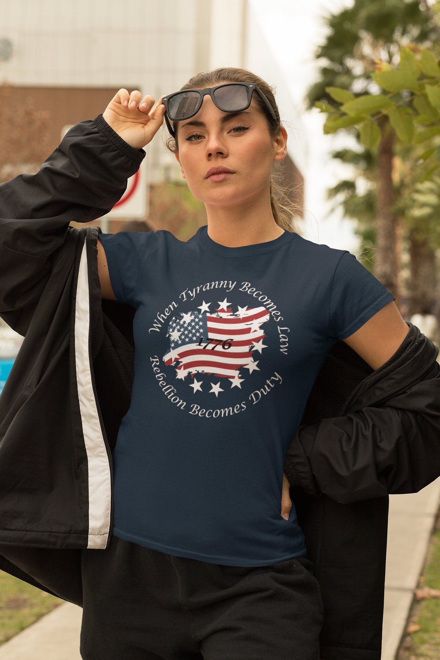 Confident young woman wearing a navy blue patriotic t-shirt with a distressed American flag and stars design, featuring the quote &#39;When Tyranny Becomes Law, Rebellion Becomes Duty - 1776.&#39; She is lifting her sunglasses while standing outdoors on a city sidewalk, dressed in a black jacket and pants.