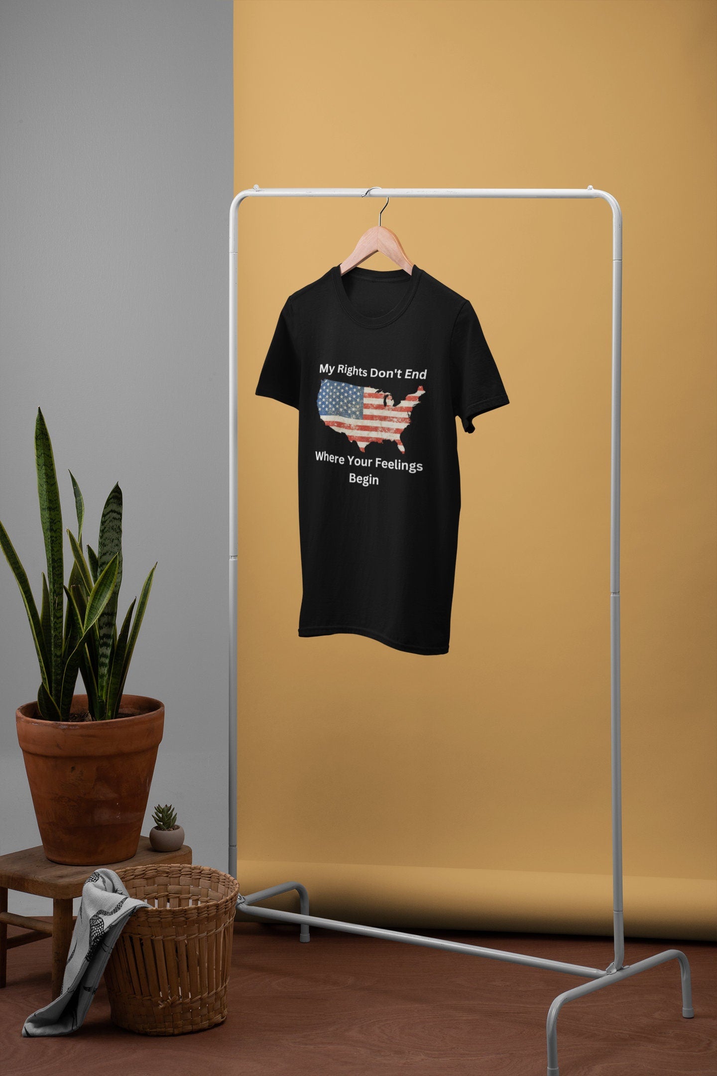 Black unisex patriotic t-shirt hanging on a clothing rack with minimalist décor, featuring a distressed American flag in the shape of the USA and the bold quote “My Rights Don’t End Where Your Feelings Begin.” Ideal for Etsy product listings targeting conservative fashion, freedom of speech apparel, and American pride shirts. Styled in a neutral studio setting with potted plants and wicker basket for a rustic, modern appeal.