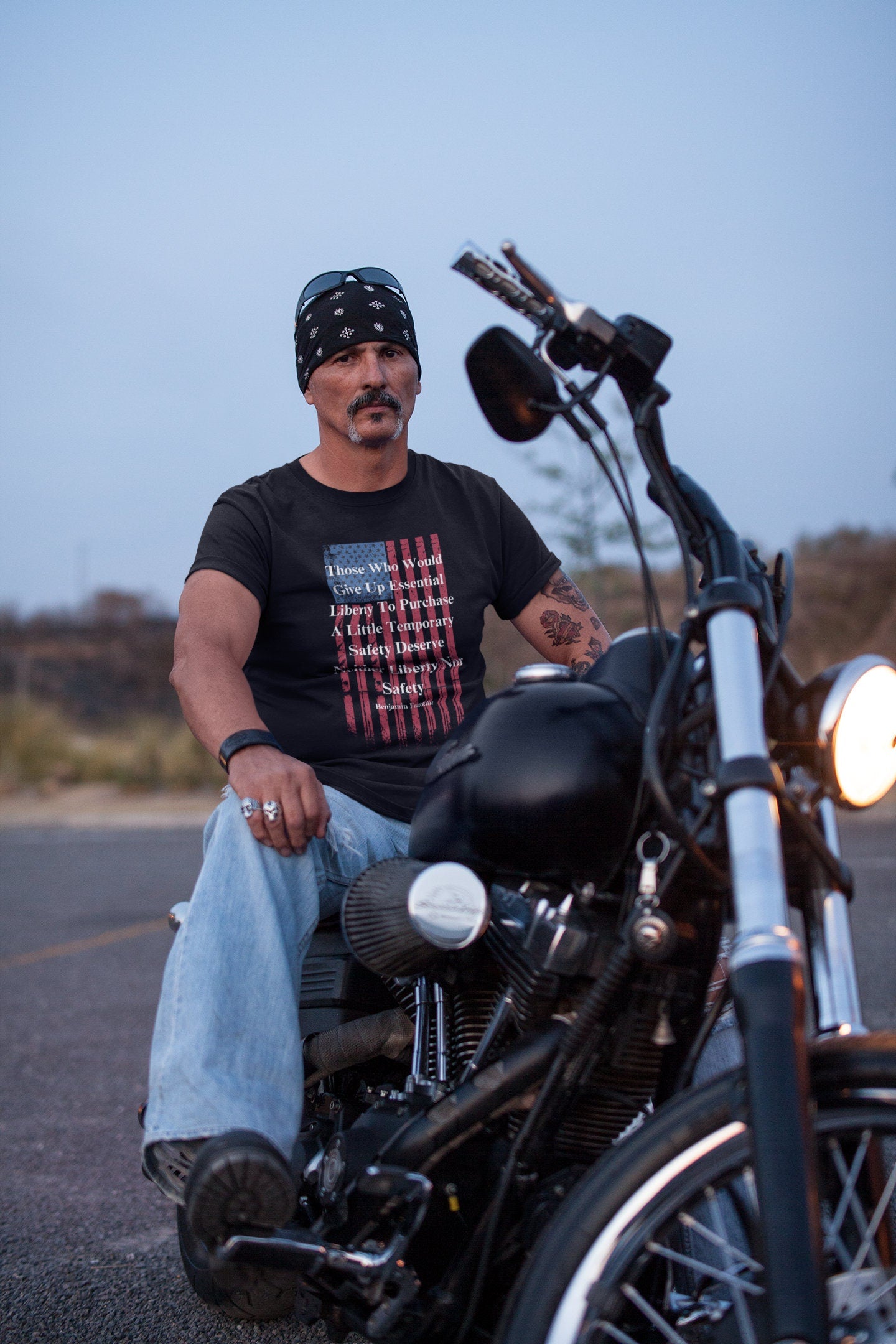 A man with a mustache and goatee sits confidently on a black motorcycle in an outdoor setting at dusk. He wears a black bandana, sunglasses resting on his head, and a black graphic T-shirt featuring a distressed American flag and a quote by Benjamin Franklin: “Those Who Would Give Up Essential Liberty To Purchase A Little Temporary Safety Deserve Neither Liberty Nor Safety.” He has tattoos on his arm and is dressed in light blue jeans with rings and a leather wrist cuff, giving off biker look.