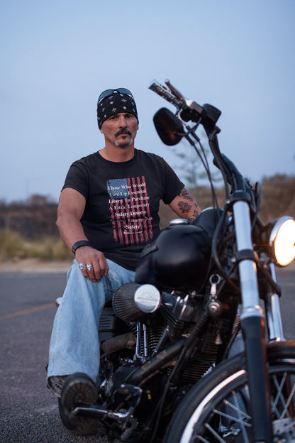 A man with a mustache and goatee sits confidently on a black motorcycle in an outdoor setting at dusk. He wears a black bandana, sunglasses resting on his head, and a black graphic T-shirt featuring a distressed American flag and a quote by Benjamin Franklin: “Those Who Would Give Up Essential Liberty To Purchase A Little Temporary Safety Deserve Neither Liberty Nor Safety.” He has tattoos on his arm and is dressed in light blue jeans with rings and a leather wrist cuff, giving off biker look.