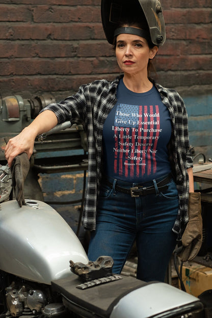 A confident woman in a workshop leans on a motorcycle while wearing welding gloves and a helmet lifted above her head. She is dressed in a plaid shirt over a navy blue graphic T-shirt that features a distressed American flag and a quote by Benjamin Franklin: “Those Who Would Give Up Essential Liberty To Purchase A Little Temporary Safety Deserve Neither Liberty Nor Safety.” The background shows industrial tools and a brick wall.