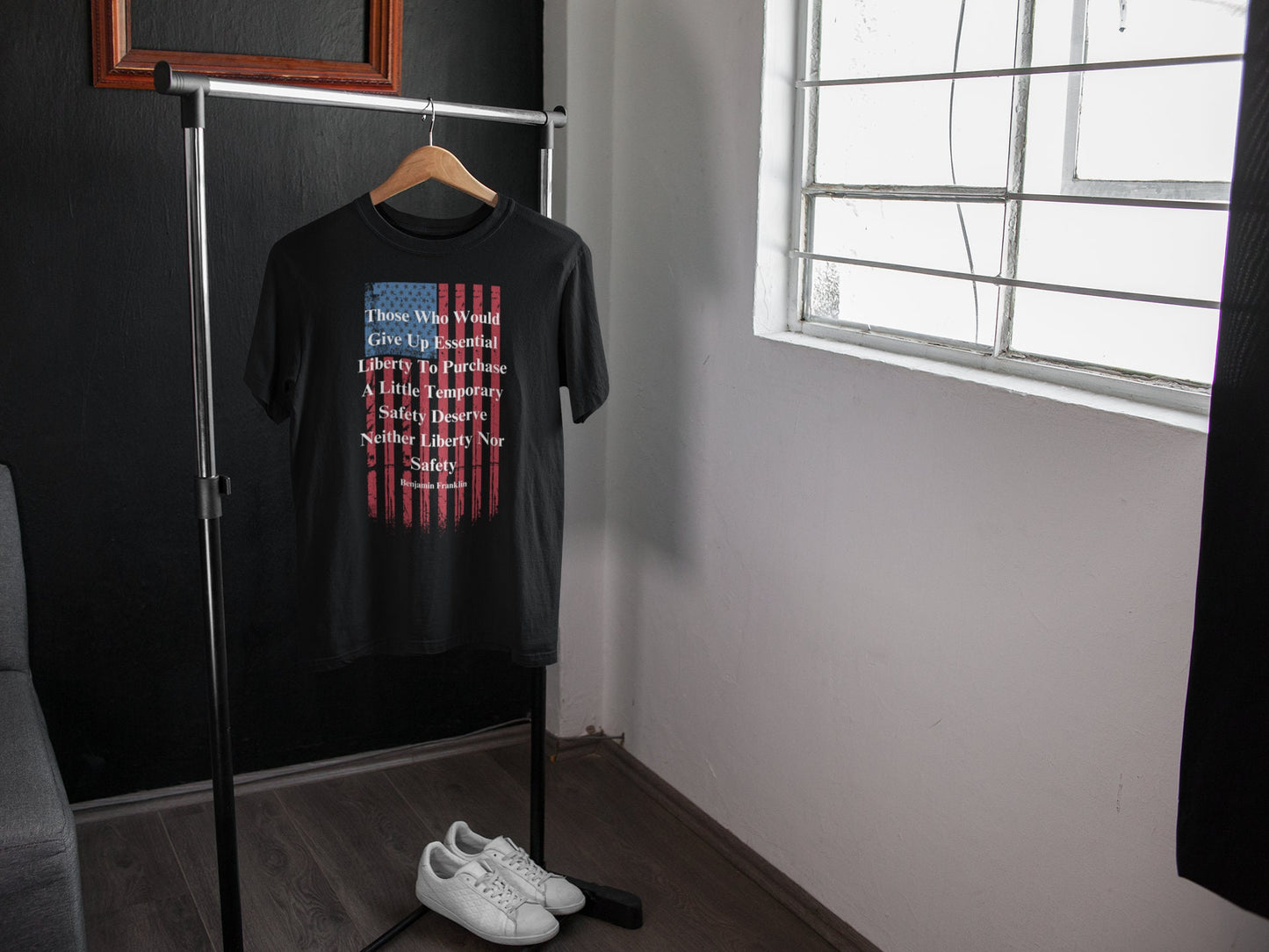 A black T-shirt hangs on a wooden hanger from a minimalist metal clothing rack in a modern, well-lit room. The shirt features a distressed American flag design and a quote by Benjamin Franklin that reads: “Those Who Would Give Up Essential Liberty To Purchase A Little Temporary Safety Deserve Neither Liberty Nor Safety.” Below the rack sits a pair of clean white sneakers on a dark wooden floor, with a window on the right allowing natural light to brighten the space.