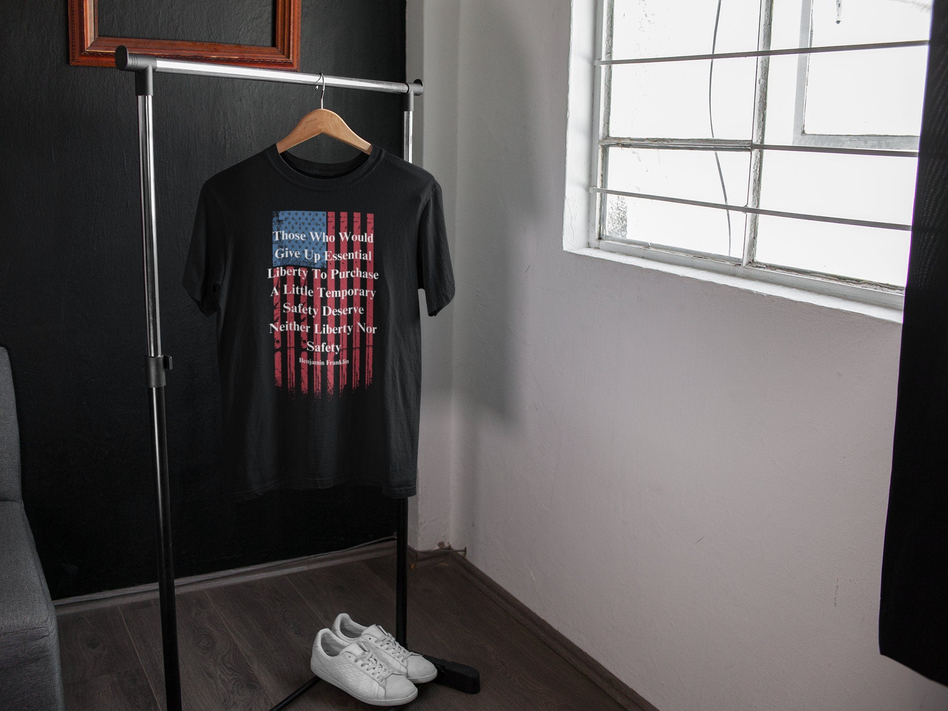 A black T-shirt hangs on a wooden hanger from a minimalist metal clothing rack in a modern, well-lit room. The shirt features a distressed American flag design and a quote by Benjamin Franklin that reads: “Those Who Would Give Up Essential Liberty To Purchase A Little Temporary Safety Deserve Neither Liberty Nor Safety.” Below the rack sits a pair of clean white sneakers on a dark wooden floor, with a window on the right allowing natural light to brighten the space.