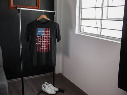 A black T-shirt hangs on a wooden hanger from a minimalist metal clothing rack in a modern, well-lit room. The shirt features a distressed American flag design and a quote by Benjamin Franklin that reads: “Those Who Would Give Up Essential Liberty To Purchase A Little Temporary Safety Deserve Neither Liberty Nor Safety.” Below the rack sits a pair of clean white sneakers on a dark wooden floor, with a window on the right allowing natural light to brighten the space.