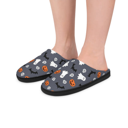 Close-up of feet wearing dark gray Halloween-themed slippers featuring a playful pattern of white ghosts, black bats, orange jack-o&#39;-lanterns, and small skulls. The slippers have a black sole and are worn indoors against a white background.