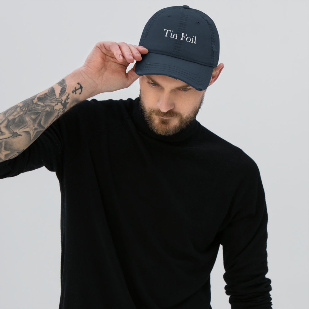 A man wearing a navy blue baseball cap with the words &quot;Tin Foil&quot; embroidered in white on the front. He is dressed in a black long-sleeve shirt and is tilting the brim of the cap downward with one hand. The man has a beard and a visible tattoo sleeve on his left arm. The background is a plain light gray, keeping the focus on the cap and the model.