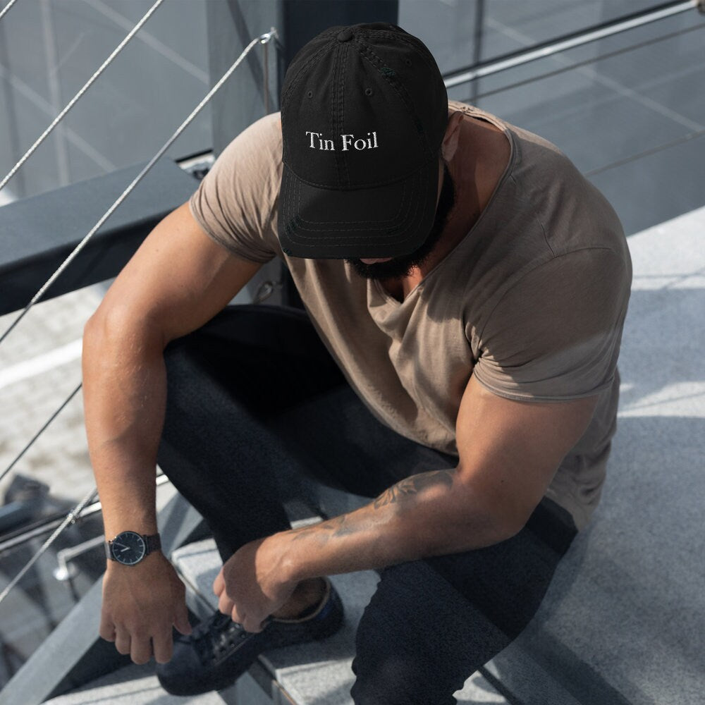 A man wearing a black distressed baseball cap with the words &quot;Tin Foil&quot; embroidered in white on the front sits on an outdoor staircase. He is dressed in a taupe t-shirt, dark pants, and black sneakers, with a black watch on his wrist. The setting appears modern and industrial, with metal railings and concrete steps, and the man is looking downward with his arms resting on his knees.