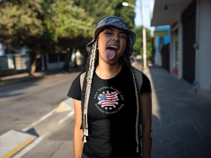 Playful young woman with long braided hair wearing a black t-shirt featuring a distressed American flag and stars design with the quote &#39;When Tyranny Becomes Law, Rebellion Becomes Duty - 1776.&#39; She sticks out her tongue and smiles while standing on a sunny urban street, wearing a patterned bucket hat and backpack.