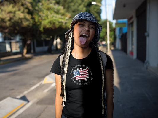 Playful young woman with long braided hair wearing a black t-shirt featuring a distressed American flag and stars design with the quote &#39;When Tyranny Becomes Law, Rebellion Becomes Duty - 1776.&#39; She sticks out her tongue and smiles while standing on a sunny urban street, wearing a patterned bucket hat and backpack.