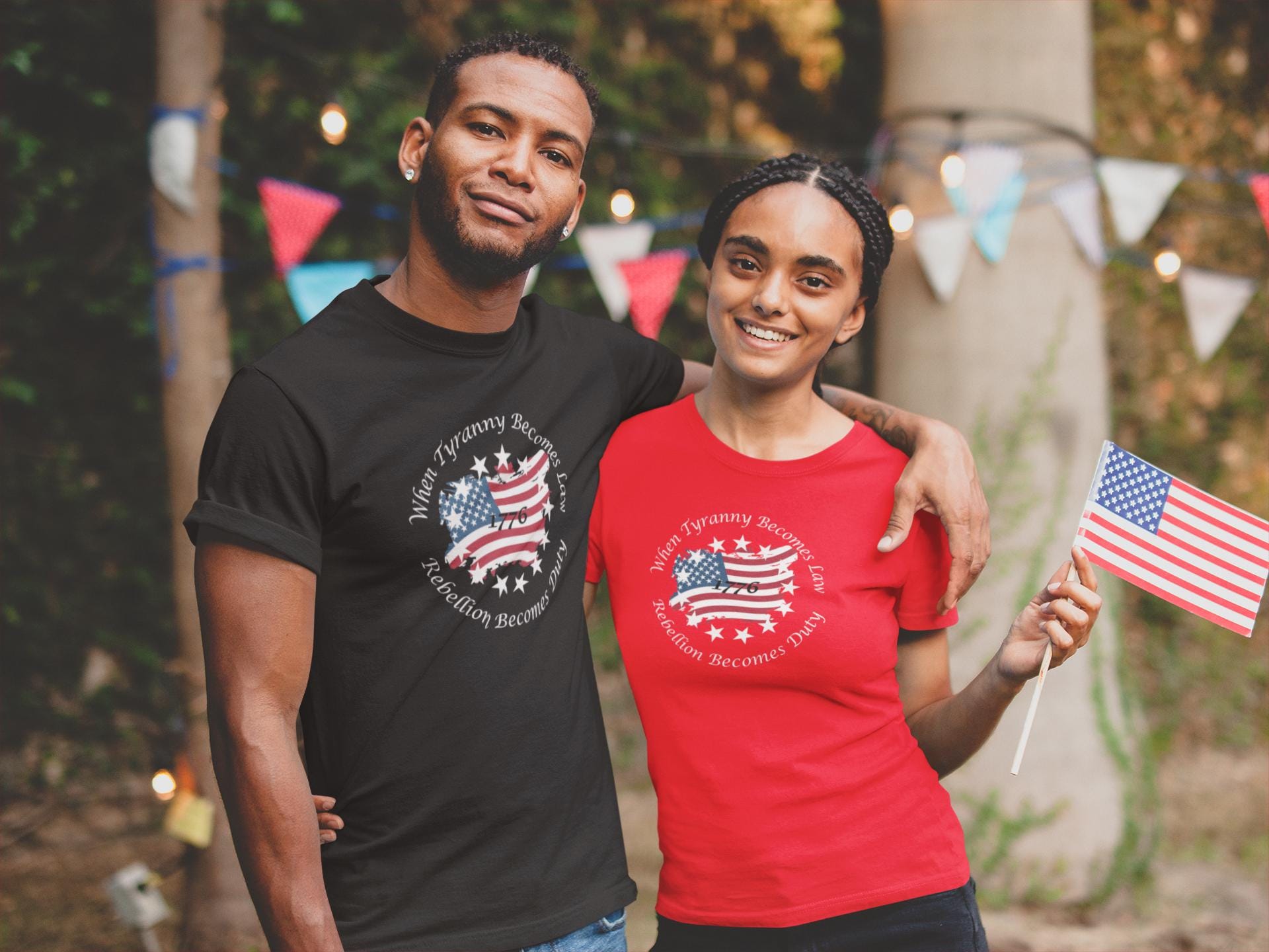 Smiling couple celebrating outdoors, wearing patriotic t-shirts with a distressed American flag and stars design featuring the quote &#39;When Tyranny Becomes Law, Rebellion Becomes Duty - 1776.&#39; The man wears a black shirt, while the woman wears a red one and holds a small American flag. Colorful bunting and string lights decorate the festive background.