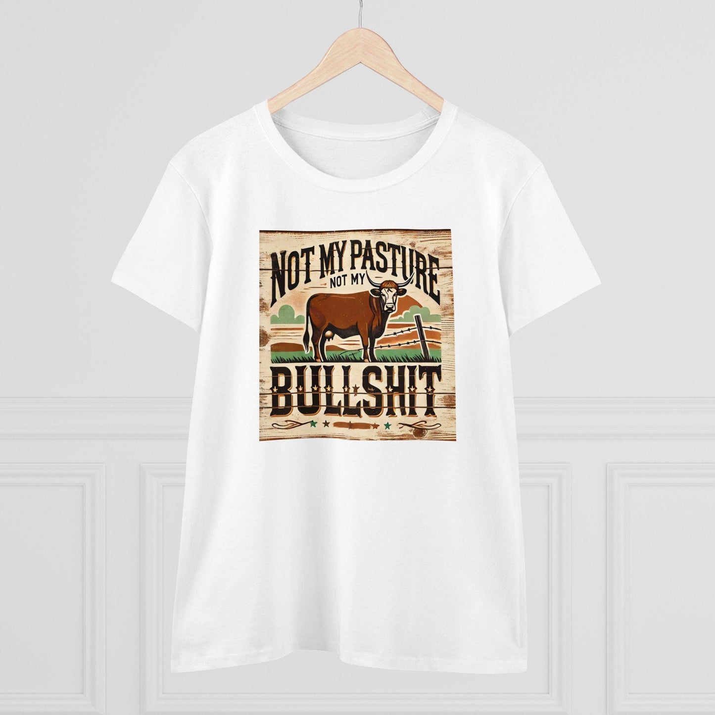 A white T-shirt hangs on a wooden hanger against a light-colored wall. The shirt features a rustic, western-style graphic of a brown cow standing in a field with a broken fence and stylized background. Bold text above and below the image reads: &quot;Not my pasture, not my bullshit.&quot; The design has a distressed wood texture for a vintage farmhouse look.