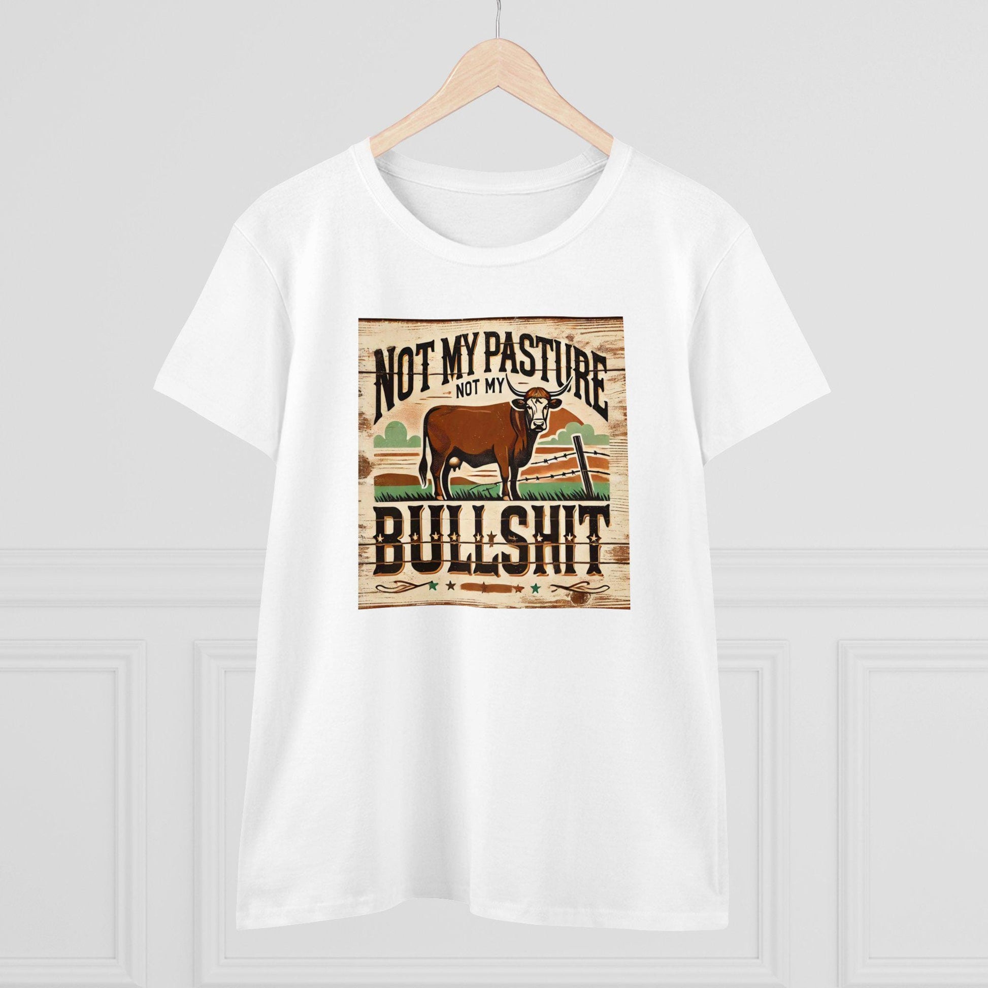 A white T-shirt hangs on a wooden hanger against a light-colored wall. The shirt features a rustic, western-style graphic of a brown cow standing in a field with a broken fence and stylized background. Bold text above and below the image reads: &quot;Not my pasture, not my bullshit.&quot; The design has a distressed wood texture for a vintage farmhouse look.