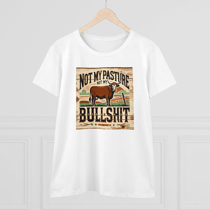 A white T-shirt hangs on a wooden hanger against a light-colored wall. The shirt features a rustic, western-style graphic of a brown cow standing in a field with a broken fence and stylized background. Bold text above and below the image reads: &quot;Not my pasture, not my bullshit.&quot; The design has a distressed wood texture for a vintage farmhouse look.