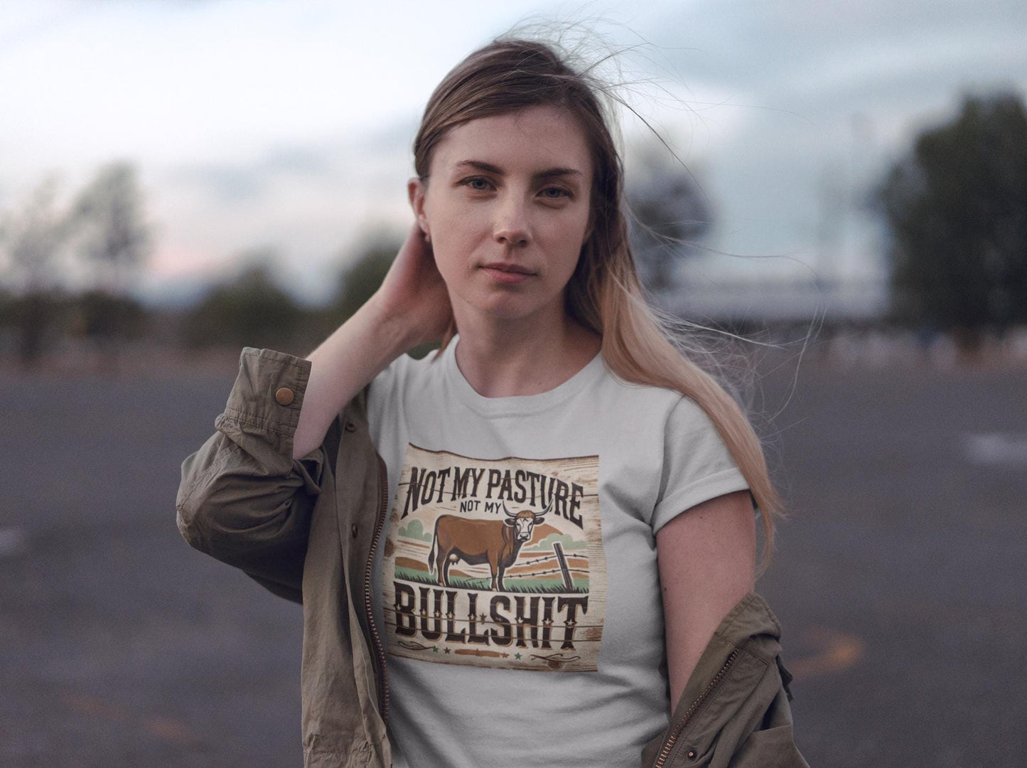 A woman stands outdoors in a parking lot at dusk, with a calm expression and one hand touching her neck. She is wearing a light-colored T-shirt with a rustic, western-style graphic featuring a cow and the bold phrase: &quot;Not my pasture, not my bullshit.&quot; An olive green jacket hangs loosely off her shoulders, and the wind gently blows her hair as the sky transitions to evening.