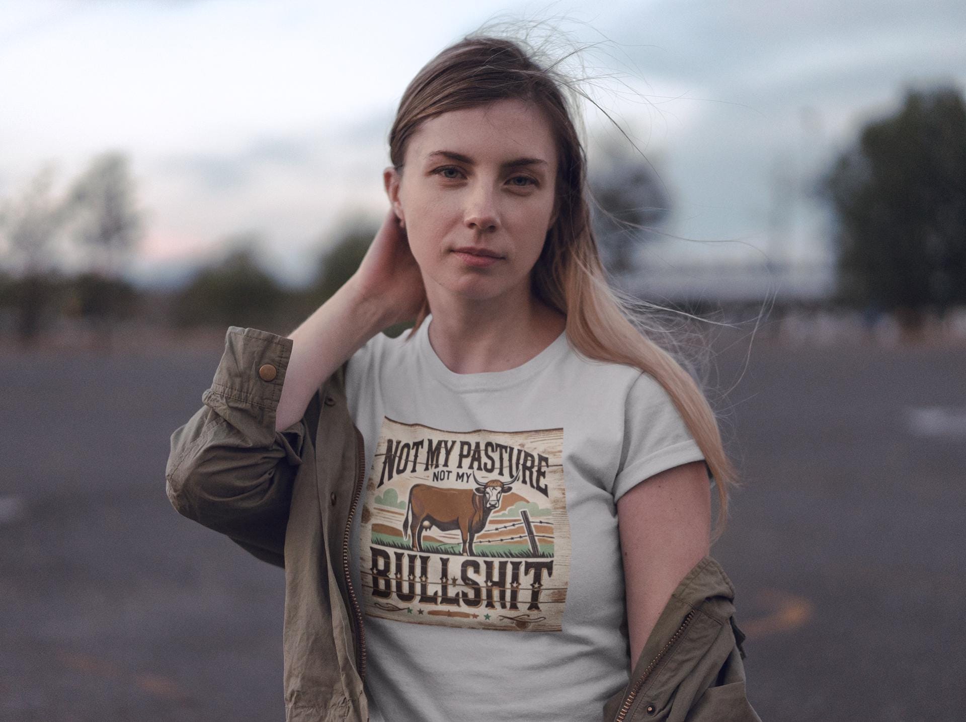 A woman stands outdoors in a parking lot at dusk, with a calm expression and one hand touching her neck. She is wearing a light-colored T-shirt with a rustic, western-style graphic featuring a cow and the bold phrase: &quot;Not my pasture, not my bullshit.&quot; An olive green jacket hangs loosely off her shoulders, and the wind gently blows her hair as the sky transitions to evening.