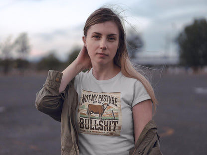 A woman stands outdoors in a parking lot at dusk, with a calm expression and one hand touching her neck. She is wearing a light-colored T-shirt with a rustic, western-style graphic featuring a cow and the bold phrase: &quot;Not my pasture, not my bullshit.&quot; An olive green jacket hangs loosely off her shoulders, and the wind gently blows her hair as the sky transitions to evening.