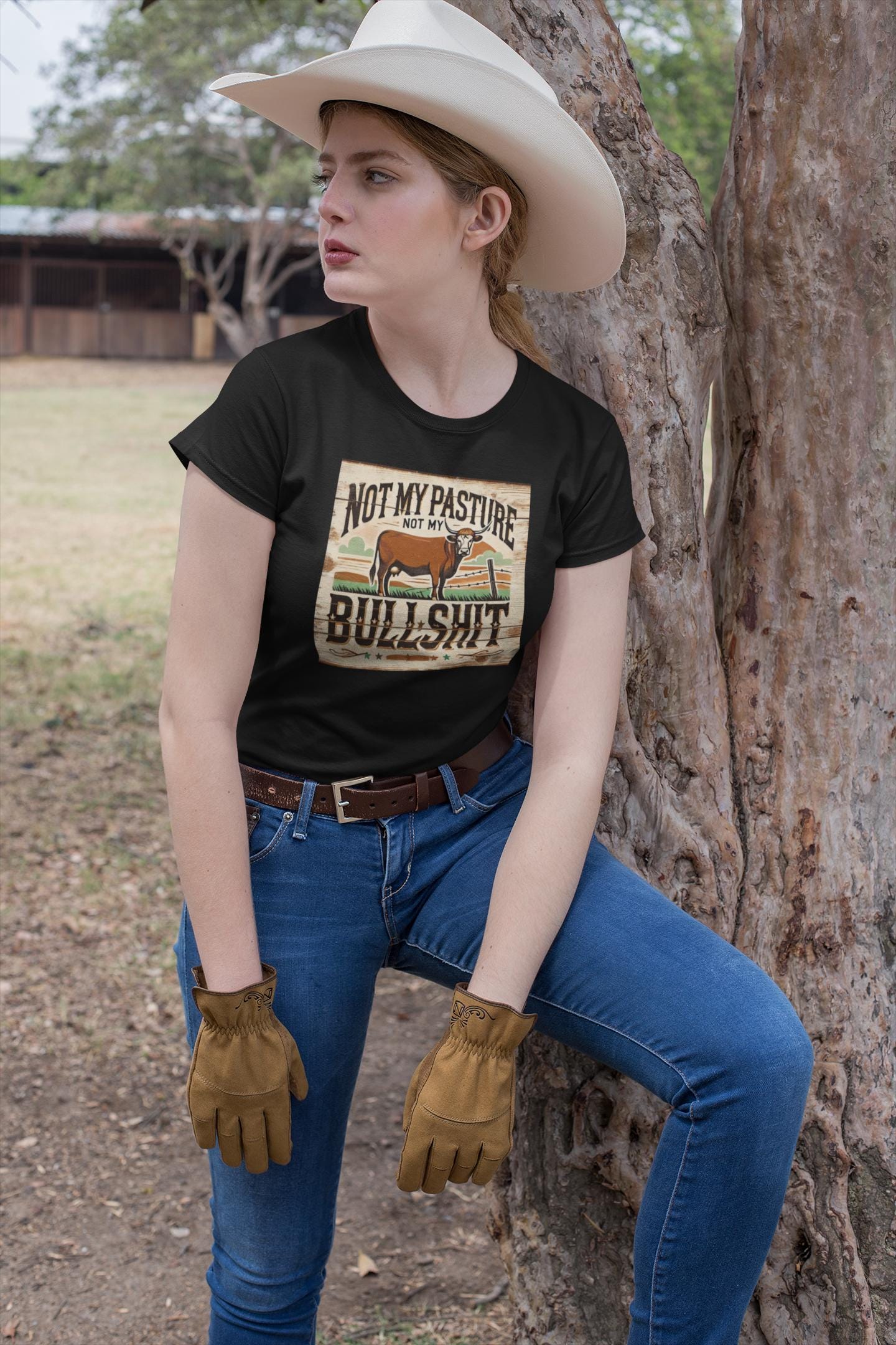 A woman wearing a white cowboy hat, tan work gloves, and blue jeans leans against a tree in a rural setting. She is dressed in a black T-shirt featuring a vintage-style graphic of a cow and a western landscape, with bold text that reads: &quot;Not my pasture, not my bullshit.&quot; She gazes off to the side with a serious expression, conveying a confident, no-nonsense attitude.