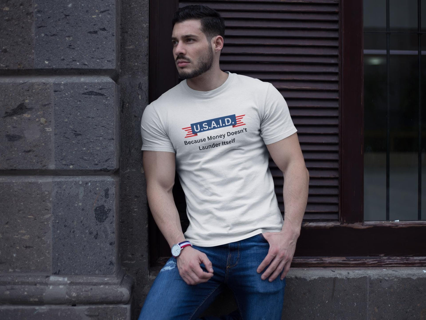A man leans casually against a stone wall outside a building, wearing a fitted white T-shirt and blue jeans. The T-shirt features a parody logo styled like a government agency emblem, reading: &quot;U.S.A.I.D. — Because Money Doesn’t Launder Itself&quot; in bold text. He has a trimmed beard, wears a watch, and gazes off to the side with a serious expression. The urban setting and lighting create a modern, street-style vibe.