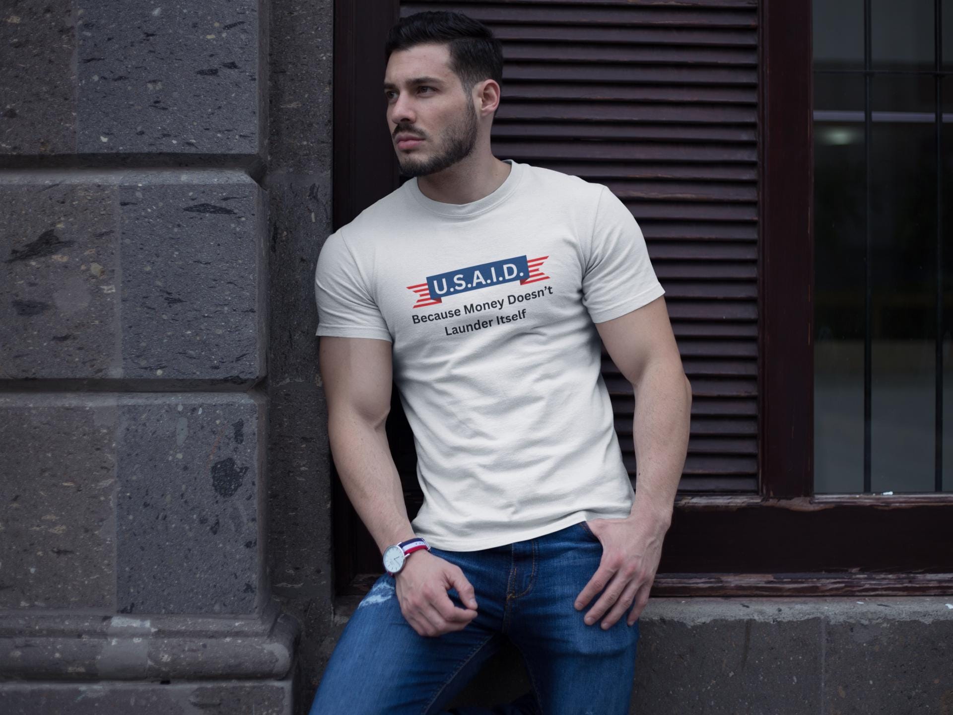 A man leans casually against a stone wall outside a building, wearing a fitted white T-shirt and blue jeans. The T-shirt features a parody logo styled like a government agency emblem, reading: &quot;U.S.A.I.D. — Because Money Doesn’t Launder Itself&quot; in bold text. He has a trimmed beard, wears a watch, and gazes off to the side with a serious expression. The urban setting and lighting create a modern, street-style vibe.