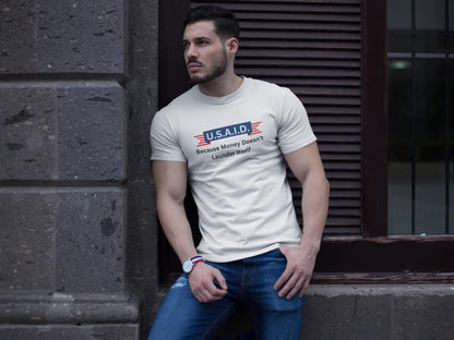 A man leans casually against a stone wall outside a building, wearing a fitted white T-shirt and blue jeans. The T-shirt features a parody logo styled like a government agency emblem, reading: &quot;U.S.A.I.D. — Because Money Doesn’t Launder Itself&quot; in bold text. He has a trimmed beard, wears a watch, and gazes off to the side with a serious expression. The urban setting and lighting create a modern, street-style vibe.