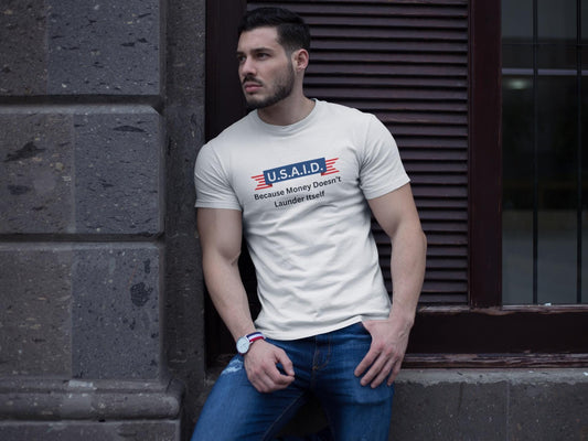 A man leans casually against a stone wall outside a building, wearing a fitted white T-shirt and blue jeans. The T-shirt features a parody logo styled like a government agency emblem, reading: &quot;U.S.A.I.D. — Because Money Doesn’t Launder Itself&quot; in bold text. He has a trimmed beard, wears a watch, and gazes off to the side with a serious expression. The urban setting and lighting create a modern, street-style vibe.