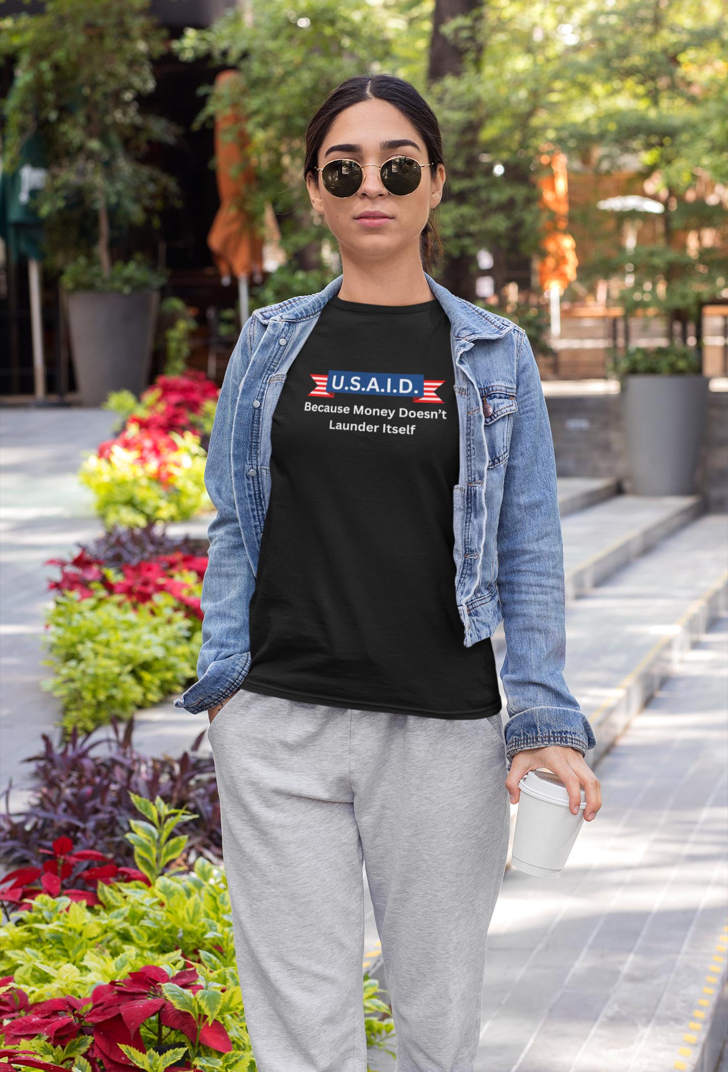 A woman stands confidently on a city sidewalk lined with colorful plants, holding a white coffee cup. She is wearing sunglasses, a denim jacket, gray sweatpants, and a black T-shirt with a parody design. The shirt features the text: &quot;U.S.A.I.D. — Because Money Doesn’t Launder Itself&quot; in bold white and red letters, mimicking a government agency logo. The overall look is casual with a touch of satire.